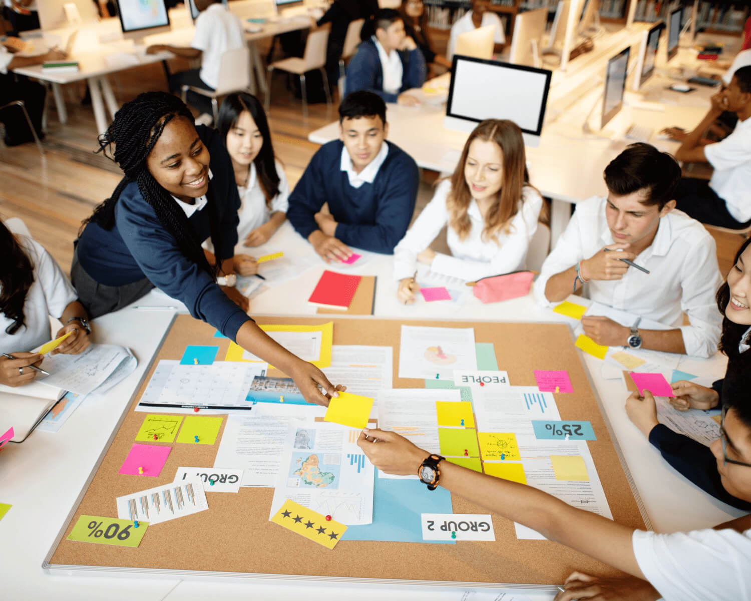 Un groupe d'étudiants est assis autour d'une table, collaborant sur un projet. Ils utilisent des notes autocollantes colorées et des documents pour organiser leurs idées sur un tableau en liège.