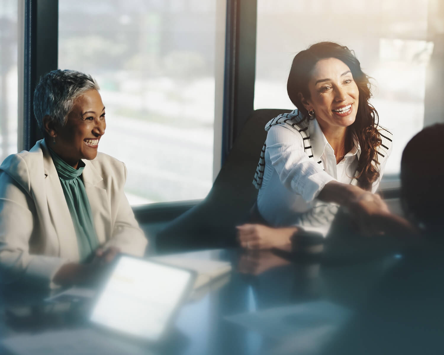 Deux femmes sont assises à une table de réunion, engagées dans une conversation. Elles semblent être dans un environnement professionnel, avec des sourires et une ambiance détendue. La lumière naturelle entre par les fenêtres derrière elles.