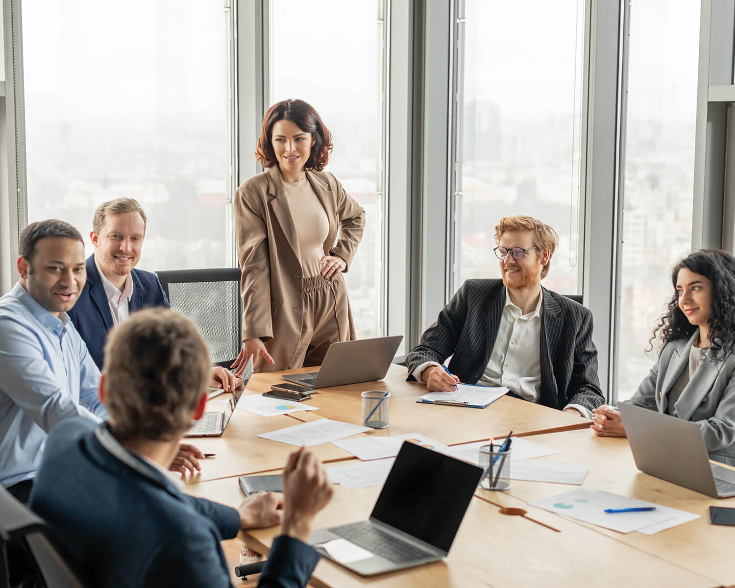 Une réunion de travail se déroule dans une salle de conférence moderne avec de grandes fenêtres. Plusieurs personnes sont assises autour d'une table, discutant et travaillant sur des ordinateurs portables. Une femme debout semble expliquer ou présenter quelque chose au groupe.